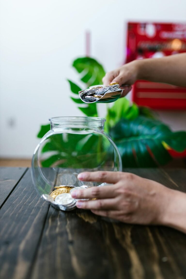 Close-up of hands placing coins into a glass jar on a wooden table with greenery in the background.